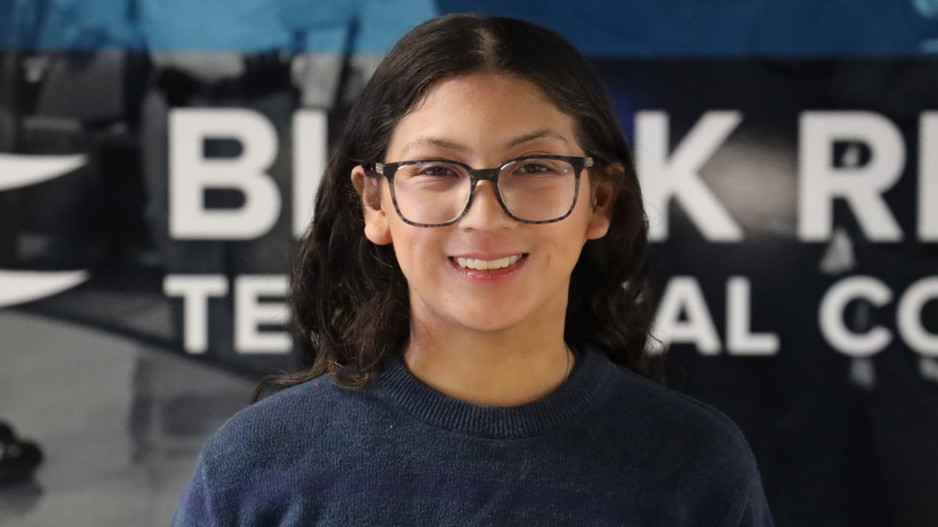 A young woman with dark curly hair and glasses smiles in front of a Black River Technical College banner.
